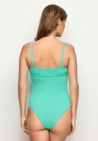 Zelené texturované jednodílné plavky s nastavitelnými tenkými ramínky a designem s vysokým vystřižením nohou, které předvádějí hladký zadní díl.