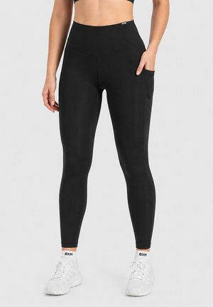 Femme portant un legging noir taille haute avec une poche latérale, des chaussettes blanches et des chaussures de sport blanches, debout devant un fond uni.