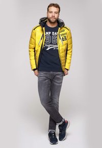 Żółta pikowana kurtka z kapturem, granatowy t-shirt z białym napisem, szare dżinsy i granatowe trampki z czerwonymi akcentami. Luźny strój.