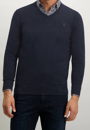 Suéter azul marino de cuello en V en tejido suave, con puños y dobladillo acanalados, y con un collar de camisa estampado visible debajo.