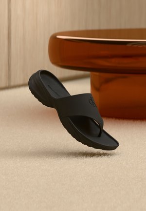 Sort slip-on sandal med hævet sål og bred rem med en tåpost, placeret på en beige tæppe nær et rundt ravfarvet bord.