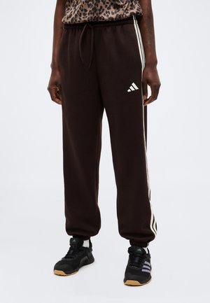 Personne portant un pantalon de survêtement Adidas noir avec des bandes blanches sur les côtés et des baskets Adidas noires avec des semelles en gomme.
