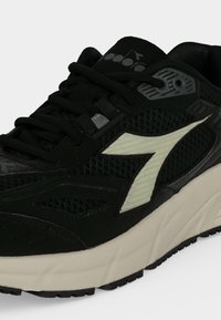 Chaussure de sport noire avec tige en mesh, logo latéral blanc, semelle épaisse beige et lacets noirs, montrée en gros plan.
