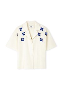 Camisa de manga corta, con botones, en lino color blanco roto con bordado floral azul en los hombros. Presenta un cuello clásico y un corte holgado.