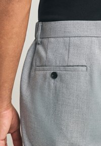 Pantaloni grigi su misura con una texture semplice, tasca posteriore con bottone nero e cinturino in vita. Vista angolata che mostra il profilo laterale.