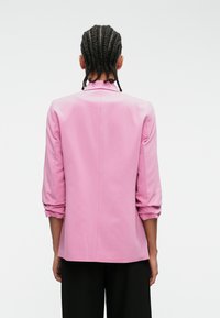 Ružový blazer so štruktúrovanými plecami, trojštvrťovými rukávmi a hladkým povrchom. Má centrálne šitie pozdĺž chrbta.