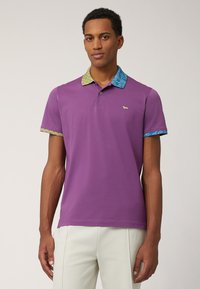 Lila polo-shirt med mönstrad krage i blått och gult, med korta ärmar och en liten logotyp på vänster sida.