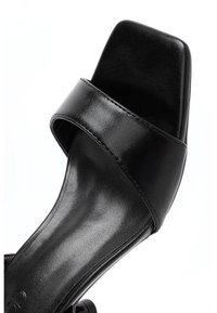 Musta neliönmuotoinen sandaali, jossa on tyylikäs, sileä nahkainen yläosa leveällä hihnalla. Pohja on kevyesti teksturoitu ilman koristeluita.