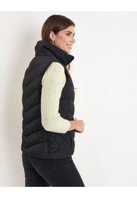 Gilet nero imbottito con colletto alto, caratterizzato da un motivo trapuntato. Indossato sopra un maglione a coste verde chiaro e jeans neri.