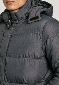 Siva puffer jakna s kapuljačom, koja se odlikuje glatkom teksturom, zatvaranjem na patentni zatvarač i velcro detaljem na ramenu za prilagodljivu prilagodbu.