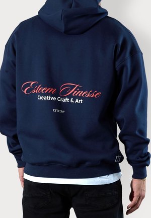 Person steht mit dem Rücken zur Kamera und trägt einen dunkelblauen Hoodie mit dem Text "Esteem Finesse Creative Craft & Art" in roten und weißen Buchstaben auf dem Rücken.