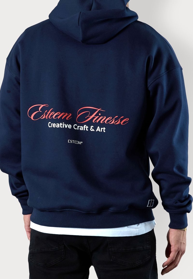 Personne de dos portant un sweat à capuche bleu marine avec le texte "Esteem Finesse Creative Craft & Art" imprimé dans le dos en lettres rouges et blanches.