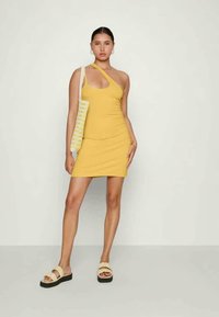 Uma mulher em um vestido mini amarelo de um só ombro, segurando uma bolsa de mão listrada e usando sandálias de plataforma bege, contra um fundo simples.