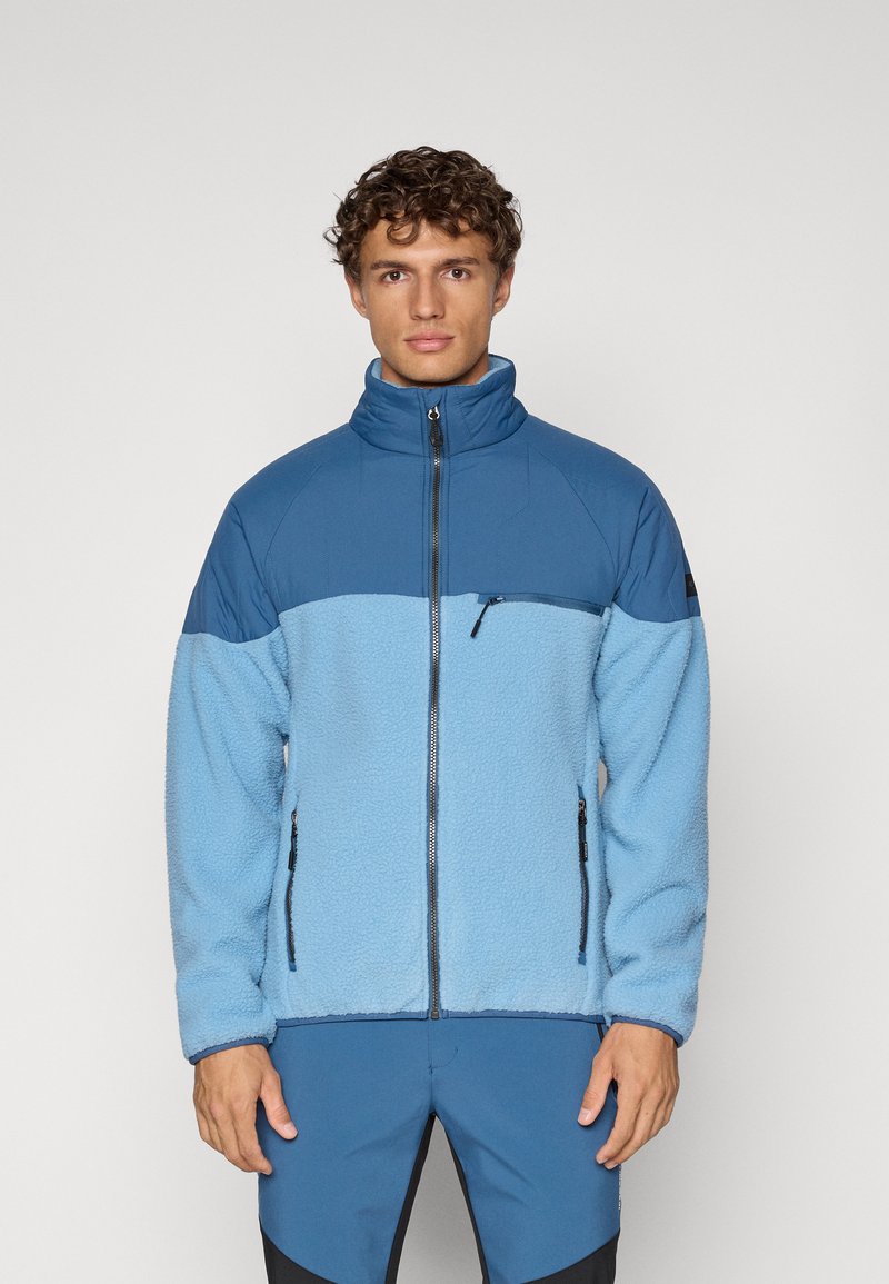 Giacca in pile azzurro chiaro con una sezione superiore blu scuro, colletto alto, zip frontale e due tasche laterali con zip; tessuto testurizzato.