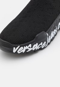 Zapatilla negra hecha de material de punto texturizado, con una suela curvada y un llamativo texto en blanco "versace jeans" a lo largo del costado.