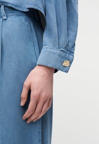 Main reposant sur le côté, portant une veste en denim bleu avec une manchette en métal et un pantalon en denim bleu taille haute assorti, sur fond blanc.