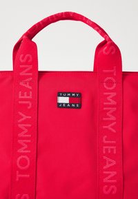Czerwona torba na ramię z grubymi uchwytami, na której wytłoczono napis "Tommy Jeans" oraz prostokątną naszywkę z logo Tommy Jeans na środku.