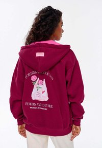 Bordový svetr s růžovým podšívkou. Na zadní straně se nachází grafika tašky naplněné růžemi s textem "Na dny, kdy potřebujete květiny a ovesné mléko."