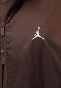 Ruskea kangastakki, jossa on korkea kaulus ja vetoketjusuljenta, valkoisella Jumpman-logolla rinnassa ja ruskeilla vetoketjun osilla.