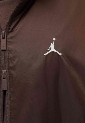 Brązowa kurtka z tkaniny z wysokim kołnierzem i zapinana na zamek, z białym logo Jumpman na piersi i brązowymi zamkami błyskawicznymi.