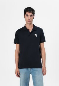 Polo majica v mornarsko modri barvi iz bombaža, z gumbnico na tri gumbe in majhnim belim logotipom na prsih. Kratki rokavi.