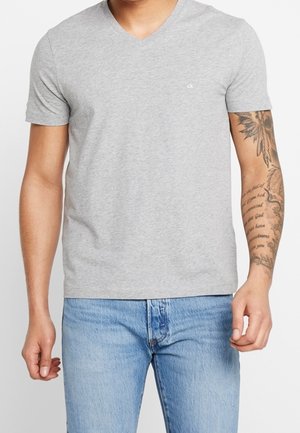 Mann trägt hellgraues V-Ausschnitt-T-Shirt mit kleinem "ck"-Logo auf der Brust und hellblaue Jeans, zeigt tätowierten rechten Unterarm.