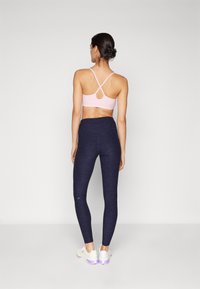 Lys rosa sports-bh med kryssede stropper, paret med mørkeblå teksturerte leggings og hvite sneakers med lilla detaljer.
