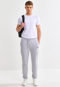 Giovane uomo in piedi, indossa una maglietta bianca, pantaloni della tuta grigi, scarpe da ginnastica bianche e porta uno zaino nero su una spalla.