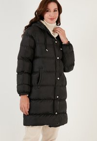 Black padded coat with a hood, featuring a zip and button closure, elastic cuffs, and two side pockets. Smooth texture and loose fit.