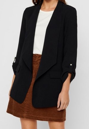 Femme portant un blazer noir ouvert sur le devant avec des manches retroussées, un haut blanc et une jupe marron en velours côtelé.