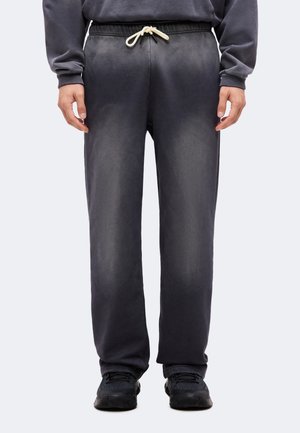 Personne portant un pantalon de survêtement gris foncé avec un cordon blanc, des baskets foncées et un sweat assorti sombre, debout les bras détendus.