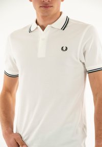 Polo bianco con tessuto testurizzato, pattina con due bottoni e logo ricamato. Presenta strisce in contrasto blu navy e verde sul colletto.
