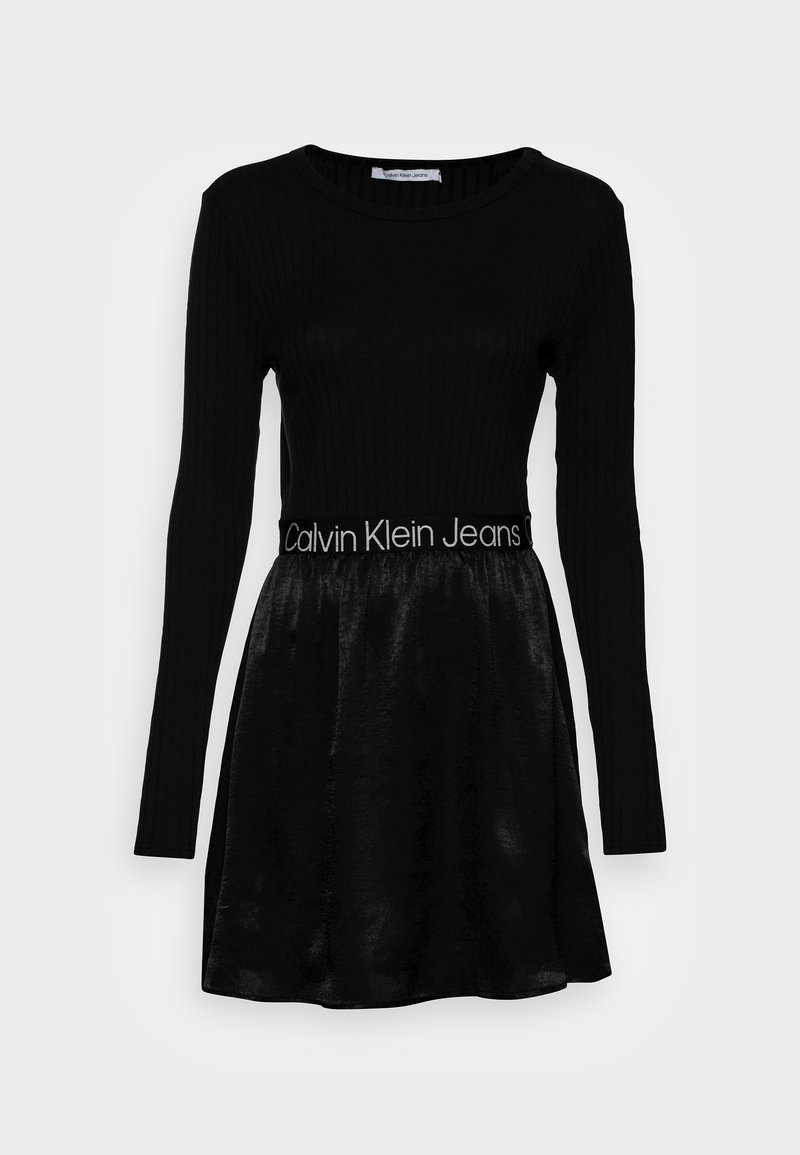 Robes noires à manches longues avec un haut côtelé, une jupe en velours et le logo Calvin Klein Jeans sur la taille, présentées sur un fond blanc.