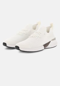 Chaussures de sport blanches avec une tige en maille, un motif texturé, des lacets plats et un accent marron au talon. Elles possèdent une semelle en caoutchouc.