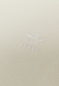 Krämfärgad tyg med en slät textur som har en vit broderad Adidas-logotyp och tre ränder ovanför.