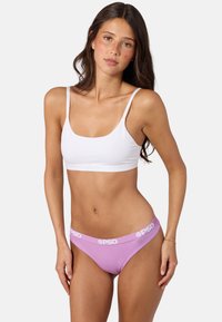 Jeune femme aux longs cheveux foncés portant un crop top blanc et des sous-vêtements lavande avec le logo PSD sur la ceinture, debout devant un fond uni.