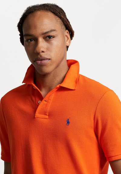 Tricou polo portocaliu cu un finisaj texturat, având un logo albastru pe partea stângă a pieptului, o deschidere cu trei nasturi și un guler pliabil.