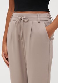 Lichtbeige broek met een trekkoord in de taille, plooien en zijzakken; gemaakt van een gladde, gestructureerde stof.