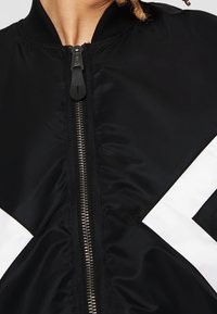 Gros plan d'une personne portant une veste noire avec une fermeture éclair argentée à l'avant et des détails à rayures diagonales blanches des deux côtés.