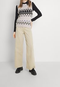 Strick-Weste mit einem hellen Lavendelgrund und geometrischen Mustern in Schwarz, Grün und Blau; kombiniert mit weiten beigen Hosen und schwarzen Schuhen.