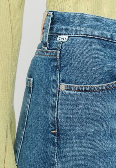 High-waisted blue denim jeans with a clean finish, featuring a small side pocket, copper-toned stitching, and a leather label near the waistband.