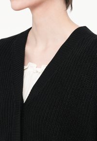 Crna pletena kardigan s V-izrezom, s rebrastom teksturom. Ispod, vidljiva je bijela bluzica s nabrane dekolteom.
