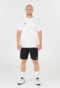 Vit polo-shirt med tre knappar, logotyp på bröstet; svarta shorts med logotyp. Modellen bär vita strumpor och svarta sportskor.