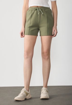 Olivengrønne shorts med en glat tekstur, elastisk talje og snoretræk. Bæres med lyse sneakers, der fremhæver et afslappet design.