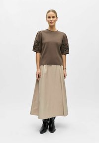 Femme debout portant un haut marron à manches courtes avec broderies florales, une longue jupe beige, des bottes noires et des boucles d'oreilles en argent, sur fond blanc.