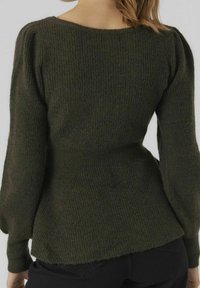 Donna che indossa un maglione a coste di un verde oliva scuro con maniche lunghe puffate e pantaloni neri, vista da dietro su uno sfondo chiaro.