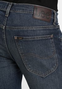 Vaqueros de mezclilla azul oscuro con superficie texturizada, que cuentan con una etiqueta de cuero marrón en la cintura y un solo bolsillo trasero con detalle de costura.