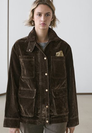 Ung person iført en mørkebrun fløjlsjakke med store lommer og et mønstret lommetørklæde, stående indendørs mod en ensfarvet baggrund.