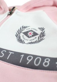Sweat  à capuche rose avec tissu blanc au niveau du col, arborant un logo avec une fleur à l'intérieur d'un losange et des accents de laurier. Le bord noir affiche "EST 1908".