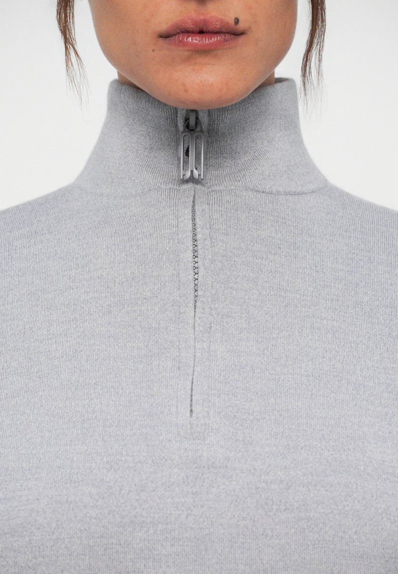 Sweater zippé gris clair avec un col haut, présentant un tissu lisse et texturé ainsi qu'un détail de fermeture éclair sur le devant.